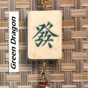 Puede incluir: Un collar con colgante que presenta una baldosa rectangular de color crema con caracteres chinos verdes, etiquetada como "Green Dragon". La baldosa est&aacute; suspendida de un cord&oacute;n marr&oacute;n y conectada a un colgante decorativo, ornamentado, rojo y dorado.