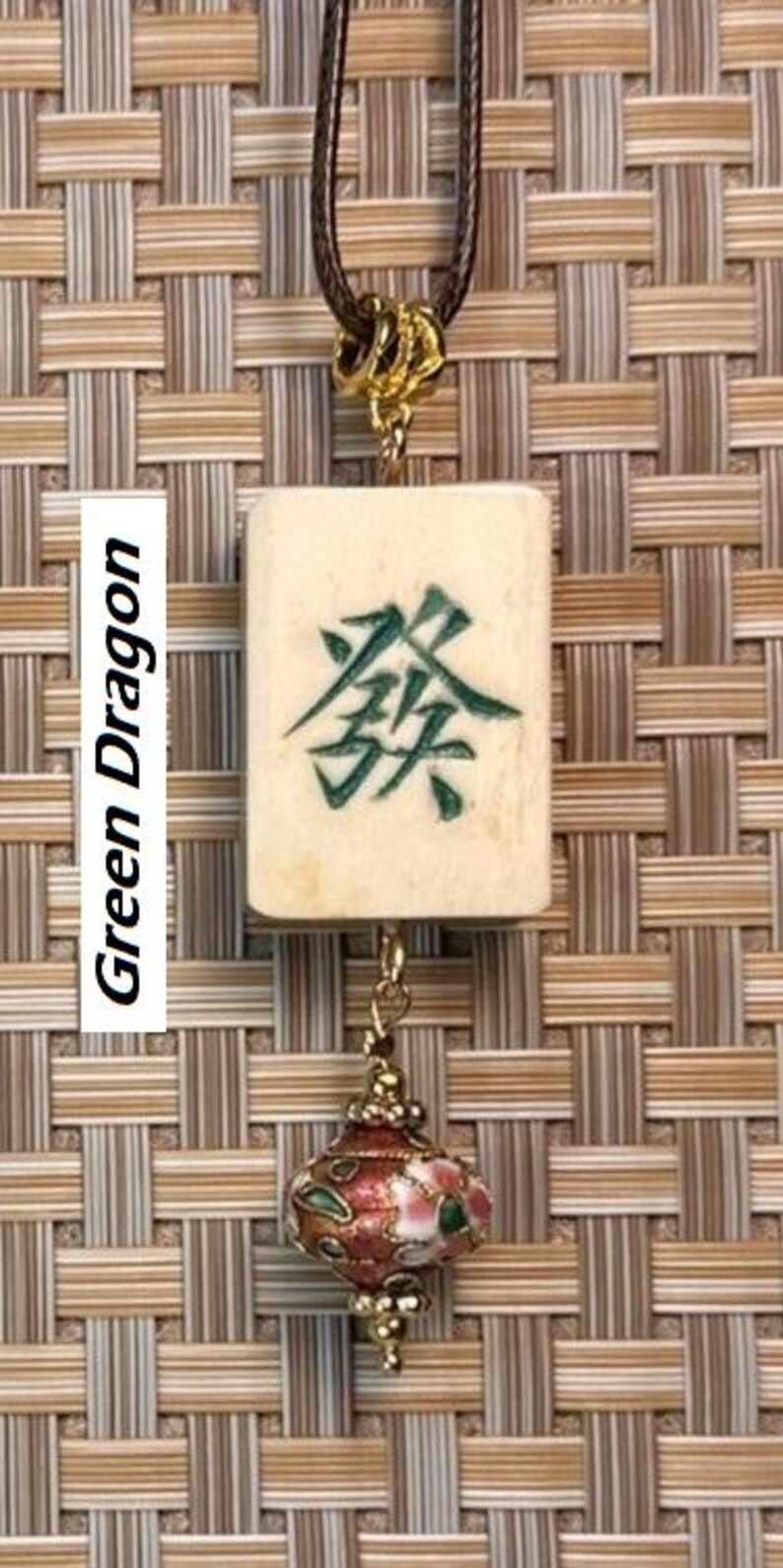 Puede incluir: Un collar con colgante que presenta una baldosa rectangular de color crema con caracteres chinos verdes, etiquetada como "Green Dragon". La baldosa est&aacute; suspendida de un cord&oacute;n marr&oacute;n y conectada a un colgante decorativo, ornamentado, rojo y dorado.