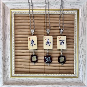 Op de afbeelding: Drie zilverkleurige kettingen, elk voorzien van een rechthoekig gebroken wit tegeltje met blauwe Chinese karakters en een rode letter E, S of W. Onder elk tegeltje hangt een donkerrode vierkante kraal met een metalen accent in het midden.