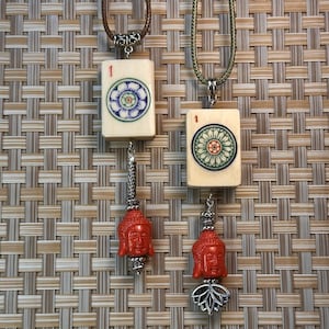 Puede incluir: Cuatro collares con colgantes. Dos presentan baldosas rectangulares color crema con diseños florales y el número "1". Los otros dos tienen colgantes de cabeza de Buda rojos. Todos están suspendidos de cordones marrones y verdes.