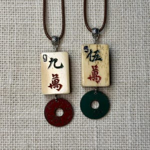 May include: Two pendant necklaces. Each features a rectangular ivory-coloured charm with Chinese characters and a circular charm. One has a red charm, the other a green charm. Both are on brown cords.
