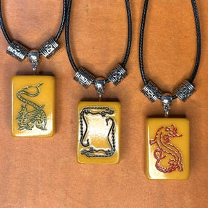 Pode incluir: Três colares com pingentes retangulares com desenhos de dragões. Os pingentes são de cor amarelo-alaranjado, e os dragões são verdes, pretos e vermelhos. Os colares têm cordões pretos e detalhes metálicos prateados.