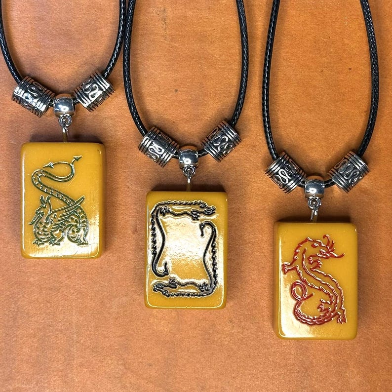 Puede incluir: Tres collares con colgantes rectangulares con dise&ntilde;os de dragones. Los colgantes son de color amarillo-naranja, y los dragones son verdes, negros y rojos. Los collares tienen cordones negros y detalles met&aacute;licos plateados.