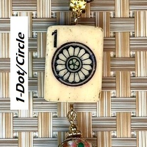 Puede incluir: Un collar con colgante que presenta un dije rectangular de color marfil con un dise&ntilde;o floral en blanco y negro y el n&uacute;mero 1. Debajo, cuelga un dije decorativo rojo, rosa y verde. El collar tiene un cord&oacute;n marr&oacute;n.