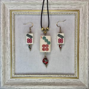 May include: A jewelry set consisting of a necklace and two earrings made from white Mahjong tiles. The necklace features a tile with the number 7 and a red patterned bead. The earrings feature smaller tiles with red and green circular designs.