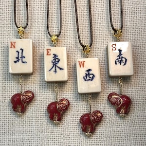 Op de afbeelding: Vier kettingen met ivoorkleurige rechthoekige hangers met blauwe Chinese karakters en de letters N, E, W en S. Elke hanger is verbonden met een rode olifantenbedel met gouden accenten. De kettingen hebben bruine koorden en goudkleurige sluitingen.
