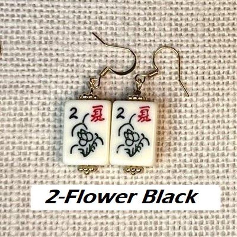 Puede incluir: Pendientes dorados con colgantes rectangulares blancos. Cada colgante muestra un n&uacute;mero 2, un car&aacute;cter chino rojo y un dise&ntilde;o floral negro. El texto "2-Flower Black" est&aacute; en la parte inferior.