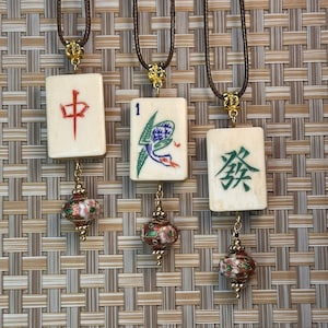Puede incluir: Tres colgantes de fichas de Mahjong de color marfil con cordones marrones. Cada ficha tiene un símbolo diferente en rojo, verde y azul. Cada colgante tiene un acento floral dorado y un pequeño dije esmaltado rojo y verde.