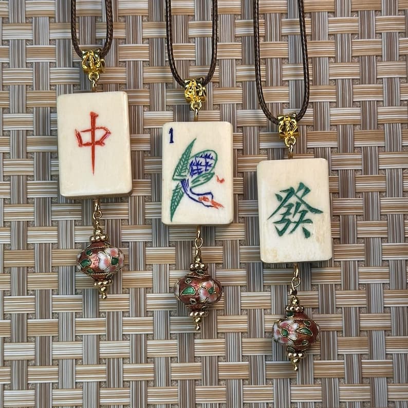 Puede incluir: Tres colgantes de fichas de Mahjong de color marfil con cordones marrones. Cada ficha tiene un s&iacute;mbolo diferente en rojo, verde y azul. Cada colgante tiene un acento floral dorado y un peque&ntilde;o dije esmaltado rojo y verde.