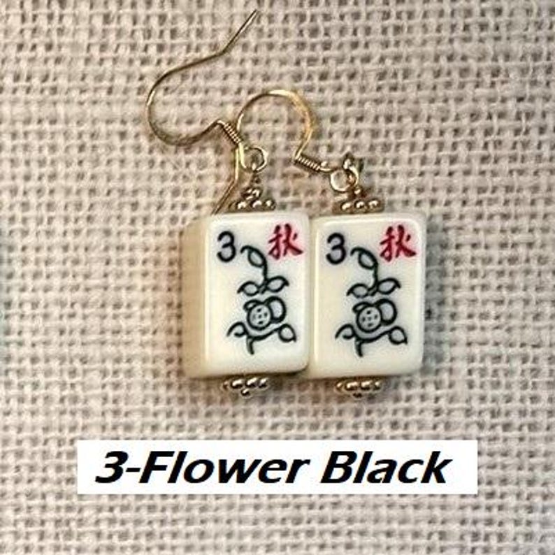 Puede incluir: Pendientes dorados con dos fichas de Mahjong de color marfil. Cada ficha muestra el n&uacute;mero 3, un car&aacute;cter rojo y un dise&ntilde;o de flor negra. El texto "3-Flower Black" es visible en la parte inferior.