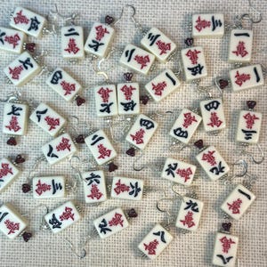 May include: A collection of ivory-colored Mahjong tile earrings with red and black Chinese characters. Each earring has a silver hook and a small, dark red bead. The earrings are scattered on a textured, light-colored surface.