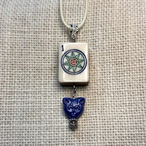 May include: A cream-colored necklace featuring a rectangular pendant with a number 1 and a circular design in blue, green, and red. Below, a blue cat-shaped charm with a silver-colored decorative base hangs from the pendant.