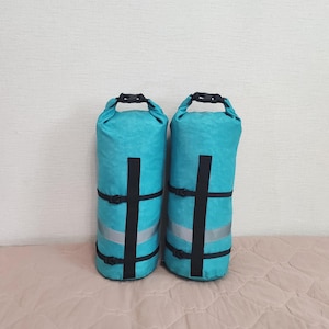 Puede incluir: Dos bolsas secas turquesas con correas negras y rayas plateadas reflectantes. Cada bolsa tiene un asa negra en la parte superior y tiene forma cilíndrica. Las bolsas están diseñadas para mantener los artículos secos.