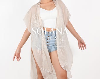 Puede incluir: Una prenda de vestir beige transparente con bordados florales blancos y un ribete de flecos. La prenda de vestir está abierta en la parte delantera y tiene mangas cortas. La modelo lleva un top blanco sin mangas y pantalones cortos de mezclilla.