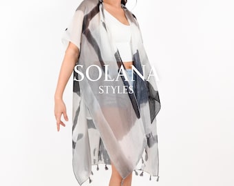 May include: A sheer white and gray kimono with black abstract designs and gray tassels. The kimono is worn over a white tank top and denim shorts. The text "SOLANA STYLES" is printed on the front of the kimono.