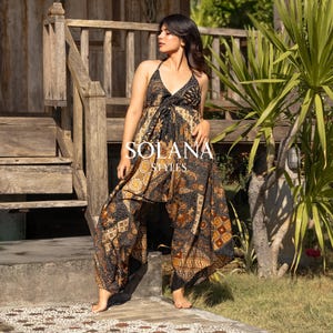 Könnte beinhalten: Ein Neckholder-Jumpsuit mit einem schwarzen, braunen und goldenen Batikmuster. Der Jumpsuit hat weite Beine und den Text "SOLANA STYLES" in Weiß. Das Model steht auf einer Steinoberfläche in der Nähe einer Holztreppe und Grün.