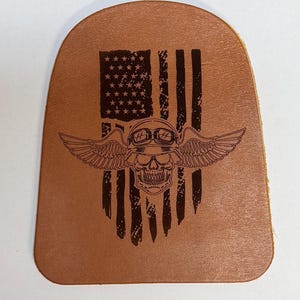 Puede incluir: Un parche de cuero color canela con borde superior redondeado y un diseño grabado con láser en marrón oscuro. El gráfico muestra una calavera con gafas y casco con alas extendidas, superpuesta sobre una bandera estadounidense desgastada.