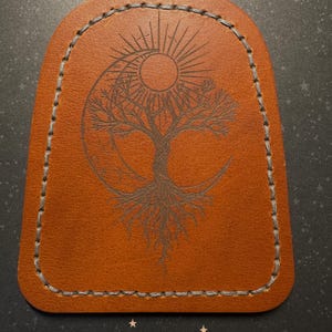Può includere: Un portachiavi in pelle marrone con un bordo cucito. Il design presenta un albero con radici, un sole e una luna crescente. L'albero è centrato nella luna e il sole è sopra l'albero. Le cuciture sono grigie.