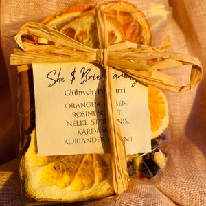 Op de afbeelding: Een doorzichtige plastic zak gevuld met gedroogde sinaasappelschijfjes, kaneelstokjes en andere specerijen. De zak is vastgebonden met een bruin lint en een label met de tekst "She & Brie" en "Glühwein Porri" met een lijst van ingrediënten in het Duits.