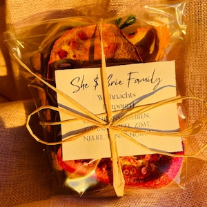 Könnte beinhalten: Eine durchsichtige Plastiktüte gefüllt mit getrockneten Fruchtscheiben, die mit einem braunen Band zusammengebunden ist. An der Tüte ist eine kleine Karte befestigt mit dem Text "She $ Brie Family Whnachts GNSOLIBEN. EFL ZIMT. NELKE, NISNUR".