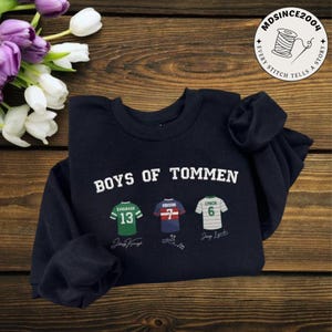 May include: Navy blue sweatshirt with the text "BOYS OF TOMMEN" in white. Below are three jersey designs with player names and numbers: Kavanagh 13, Gibson 7, and Lynch 6. The sweatshirt is displayed on a wooden surface.