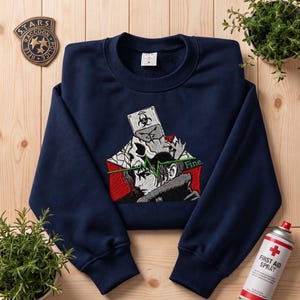 Peut inclure: Un sweat-shirt bleu marine à col rond avec un motif brodé central représentant le profil d'un personnage, un crâne et un symbole de risque biologique. Une ligne de rythme cardiaque verte traverse le motif avec le mot "Fine." en blanc.