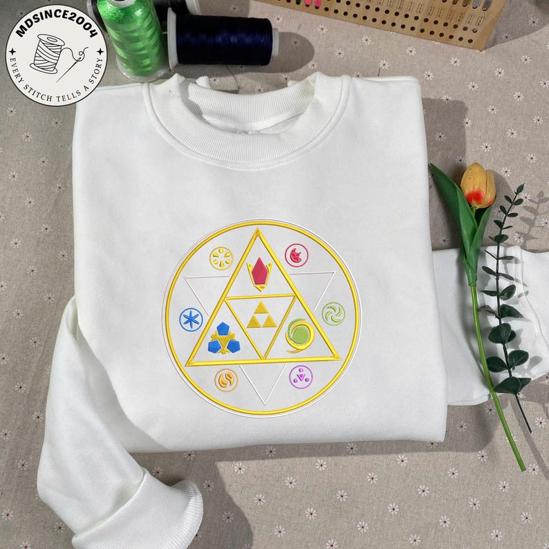 May include: White sweatshirt with a circular embroidered design featuring a gold triangle and various colorful symbols. The design is centered on the chest. The text "MDSince2004" and "Every Stitch Tells a Story" are printed on the top left.
