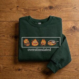 Puede incluir: Una sudadera de cuello redondo verde oscuro con un gráfico rectangular que muestra cuatro personajes de fuego animados con diversas expresiones y una sartén. Debajo del gráfico, la palabra "overstimulated" está impresa en fuente blanca.