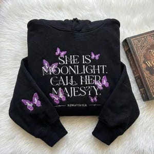 Può includere: Felpa con cappuccio nera con la scritta "SHE IS MOONLIGHT. CALL HER MAJESTY." e ricami di farfalle viola. La felpa ha anche il nome del marchio "KINGFISHER" stampato.