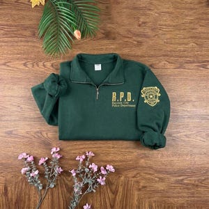 May include: A dark green quarter-zip sweatshirt featuring a gold embroidered R.P.D. Raccoon City Police Department logo on the chest and a matching gold police badge emblem on the left sleeve, with a collar and long sleeves with ribbed cuffs.