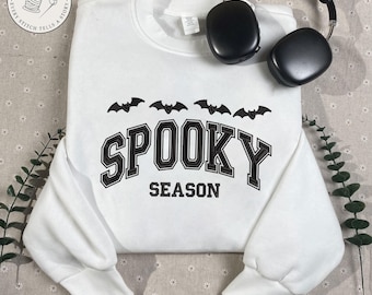 May include: White sweatshirt with the text "SPOOKY SEASON" in black, with black bat graphics above the text. Black headphones are placed on the sweatshirt. The sweatshirt has long sleeves and a crew neck.