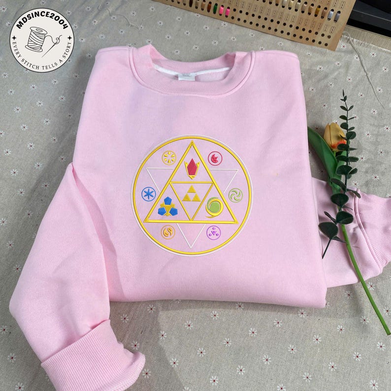 May include: A light pink sweatshirt with a crew neck and long sleeves. The front features a circular embroidered design with a gold border and various colorful geometric shapes. The design includes a triangle and other symbols.