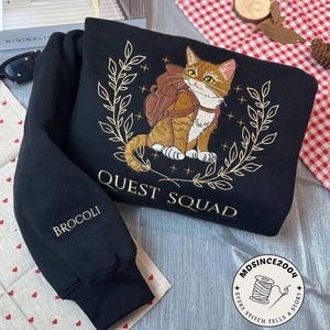 Könnte beinhalten: Schwarzer Pullover mit gesticktem Design einer Katze, die einen Rucksack trägt und von einem Kranz umgeben ist. Der Text "Quest Squad" ist unter der Katze gestickt. Der Text "Broccoli" ist auf dem linken Ärmel gestickt. Der Text "MDSINCE2004 EVERY STITCH TELLS A STORY" ist auf einem runden Patch unten rechts gestickt.