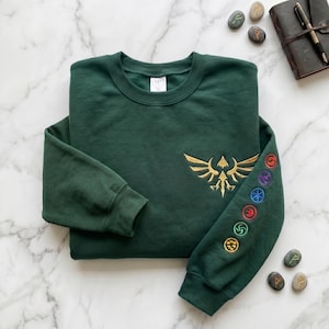 以下が含まれることがあります： 胸に金色の刺繍エンブレム、袖にカラフルな円形デザインが施された濃い緑色のスウェットシャツ。 大理石の表面に折りたたまれ、革張りのノートと石が近くにあります。