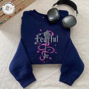 Może przedstawiać: Granatowa bluza z haftowanym srebrnym napisem "Fearful", kluczem i różowymi akcentami. Czarne słuchawki spoczywają na bluzie. Bluza ma długie rękawy.