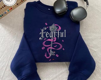 May include: A navy blue sweatshirt with the word "Fearful" embroidered in silver, along with a key and pink accents. Black headphones rest on the sweatshirt. The sweatshirt has long sleeves.