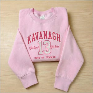 Peut inclure: Un sweat-shirt rose clair avec le nom "KAVANAGH" en lettres rouges et blanches. Le numéro "13" est en blanc avec un contour rouge. Le texte "For keeps?" et "BOYS OF TOMMEN" sont également visibles.