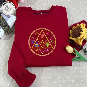 May include: A burgundy sweatshirt featuring a circular design with a yellow triangle and various symbols in yellow, blue, and green. The design is centered on the front of the sweatshirt. The cuffs and collar are ribbed.