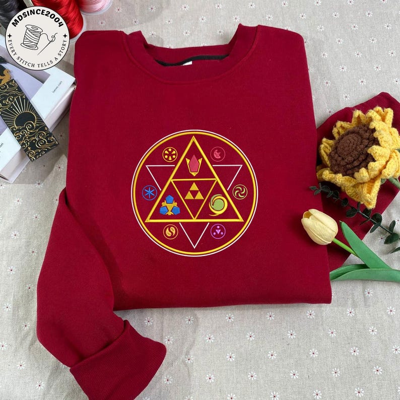 May include: A burgundy sweatshirt featuring a circular design with a yellow triangle and various symbols in yellow, blue, and green. The design is centered on the front of the sweatshirt. The cuffs and collar are ribbed.
