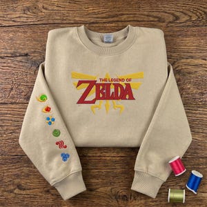 Puede incluir: Una sudadera beige con la inscripción "THE LEGEND OF ZELDA" en rojo y dorado. La manga izquierda tiene símbolos bordados. Se ven dos carretes de hilo. La sudadera está sobre una superficie de madera.