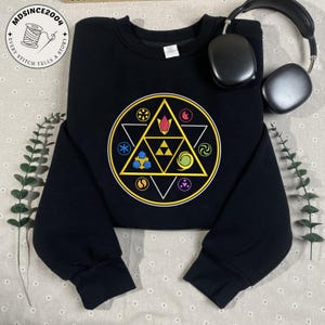 May include: Black sweatshirt featuring a colorful geometric design with a triangle and circle pattern. The design includes various symbols in yellow, blue, red, and green. A pair of black headphones rests on the top right. The sweatshirt has a crew neck.