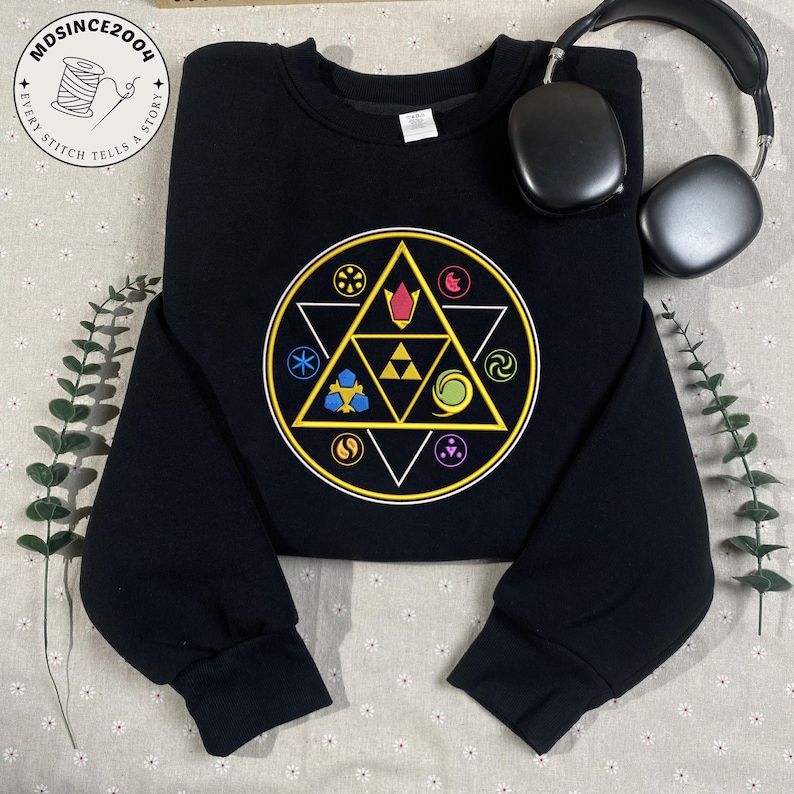 May include: Black sweatshirt featuring a colorful geometric design with a triangle and circle pattern. The design includes various symbols in yellow, blue, red, and green. A pair of black headphones rests on the top right. The sweatshirt has a crew neck.
