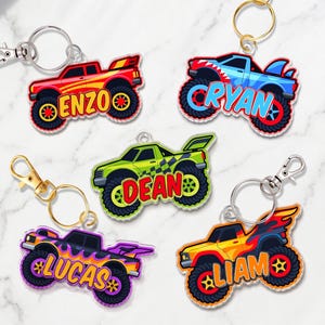 May include: Five acrylic monster truck keychains with metal lobster claw clasps. Each truck features a unique color scheme and a personalized name: ENZO in red, CRYAN in blue, DEAN in green, LUCAS in purple, and LIAM in yellow with flame graphics.