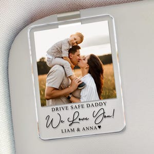 May include: A clear acrylic car visor photo holder with a family photo. The photo shows a family of three, with the text "Drive Safe Daddy, We Love You!" and names "Liam & Anna" below the photo.