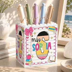 May include: A square white pen holder constructed from building blocks, featuring a lined paper pattern with the text "Miss SOPHIA" in a large letter S shaped like a pencil. The design includes colorful illustrations of bows, apples, books, and pencils, holding five pens with gold-toned clips.