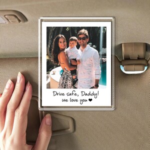 Puede incluir: Un marco de fotos de acr&iacute;lico transparente con una foto familiar adjunta a una visera de coche. La foto muestra a una familia de tres, con el texto "Drive safe, Daddy! we love you" escrito debajo.
