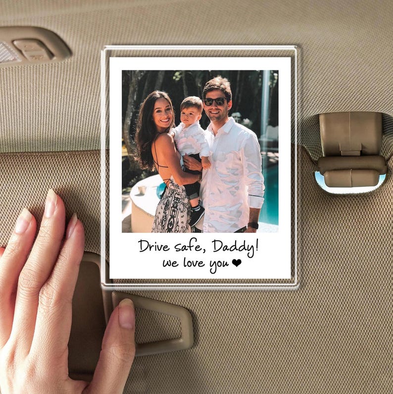 Puede incluir: Un marco de fotos de acr&iacute;lico transparente con una foto familiar adjunta a una visera de coche. La foto muestra a una familia de tres, con el texto "Drive safe, Daddy! we love you" escrito debajo.
