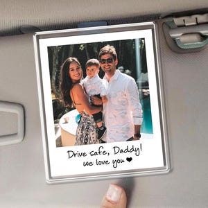 Puede incluir: Un marco de fotos rectangular transparente con un retrato familiar. La foto muestra a una mujer, un hombre y un ni&ntilde;o sonriendo. El texto en la parte inferior dice "Drive safe, Daddy! we love you ❤". El marco est&aacute; sujeto a una visera de coche.