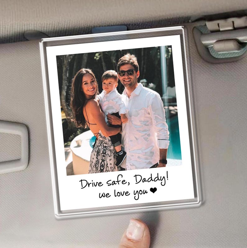 Puede incluir: Un marco de fotos rectangular transparente con un retrato familiar. La foto muestra a una mujer, un hombre y un ni&ntilde;o sonriendo. El texto en la parte inferior dice "Drive safe, Daddy! we love you ❤". El marco est&aacute; sujeto a una visera de coche.
