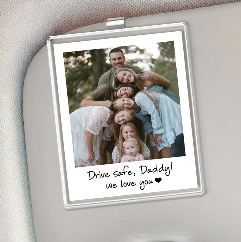 Puede incluir: Un portafotos rectangular para parasol de coche con una foto familiar. La foto muestra un grupo de personas sonriendo, con el texto "Drive safe, Daddy! we love you" en escritura negra. El soporte est&aacute; fijado a un parasol de coche.